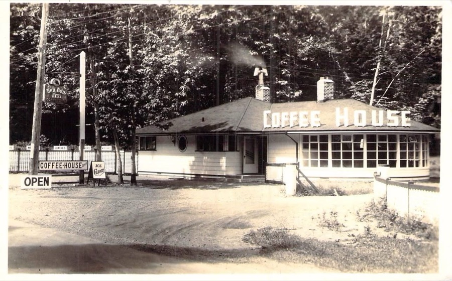 A real photo postcard of the inn's roadside coffee house: a low
                      white building with COFFEE HOUSE lettered across the roof, a curved
                      bank of windows, a sandwich board reading OPEN, and the Ben Bow Inn
                      sign hanging from a post by the highway.