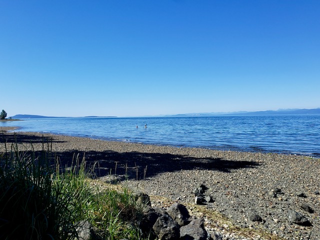A pebble and shell beach looking out over calm blue water, with two paddleboarders offshore and the mainland mountains on the horizon.